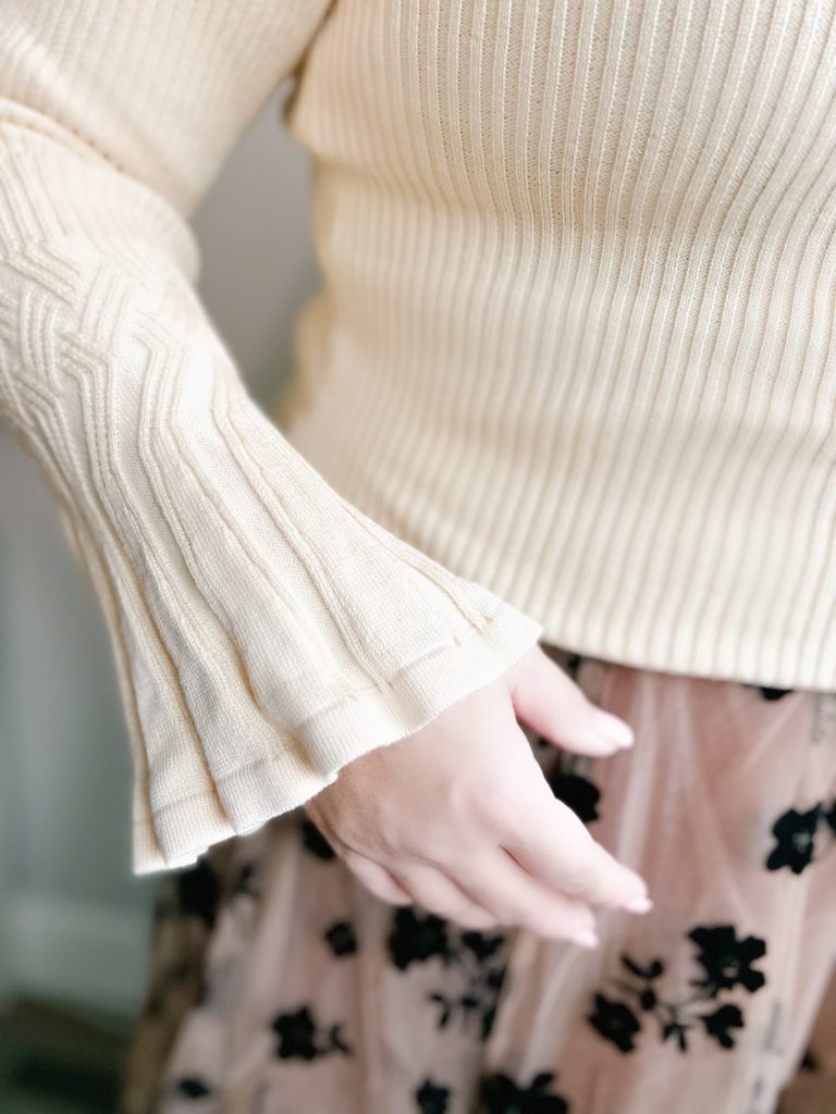 La Belle Vie Sweater in Cream with beautiful detailing at the bell cuff sleeves and at the wrists. Body of the sweater is a thin knit. Anne, an Asian woman, @PurposeInView is wearing the sweater. Camera view is from the neck down as she wears the sweater. 

