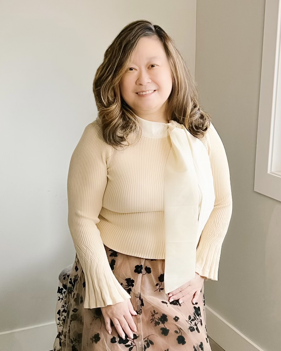 Anne, an Asian woman, is smiling at the camera. She is wearing a fine knit cream sweater with a tie detail and a champagne coloured skirt with black velvet flower overlay detail.