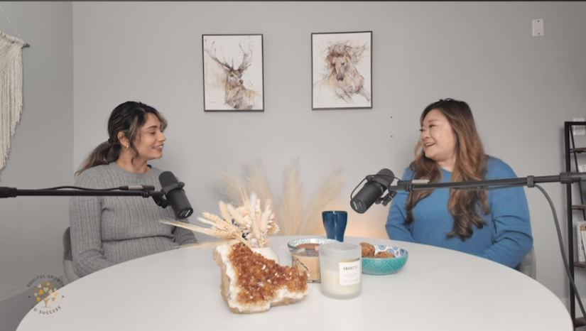 Prabhleen and Anne, an Asian woman, are sitting down at a table with podcasts microphones.